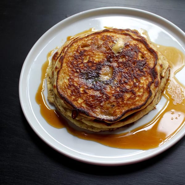 Stack of golden-brown pancakes with melting butter and maple syrup on a white plate.
